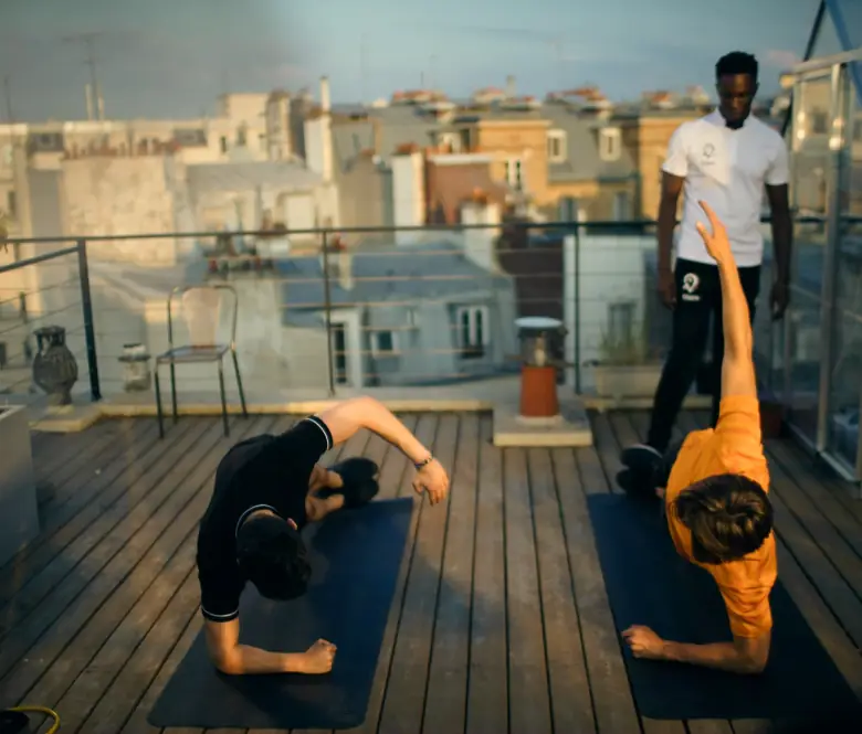 Un coach debout observe deux clients en exercice au sol, sur une terrasse au crépuscule.